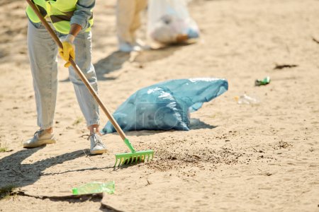Photo for A woman in a safety vest and gloves sweeps up trash on the beach, embodying the spirit of environmental stewardship and care. - Royalty Free Image