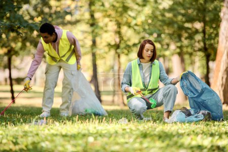 Diverse couple in safety vests and gloves stand on grassy field, actively participating in a park clean-up event.