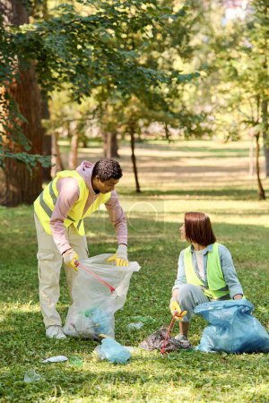 Afrikanisch-amerikanischer Mann und kaukasische Frau in Sicherheitswesten und Handschuhen arbeiten zusammen, um Müll in einem Park zu sammeln und so Umweltfreundlichkeit und Gemeinschaftspflege zu fördern.