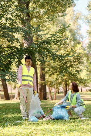 Una pareja diversa en chalecos de seguridad y guantes de pie en el césped, la limpieza de un parque juntos.
