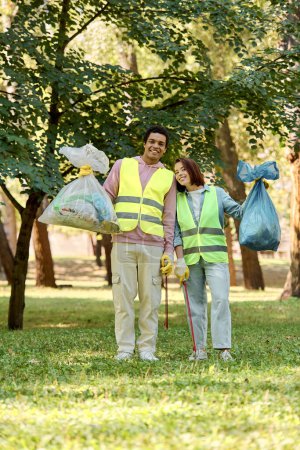 A socially active diverse loving couple in safety vests and gloves cleaning a park together, holding bags of garbage.