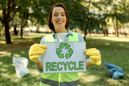 A woman wearing gloves holding a sign that says recycle, promoting environmental awareness and sustainability in a park cleanup.
