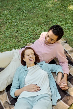 A man and woman, dressed vibrantly, lay contentedly on a blanket in the lush grass of a park, enjoying each others company.