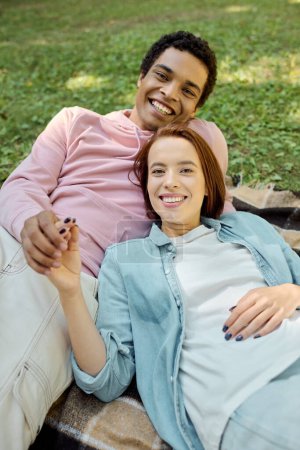 Una pareja diversa en traje vibrante se sientan juntos en un banco del parque, disfrutando de un momento pacífico y amoroso en la naturaleza.