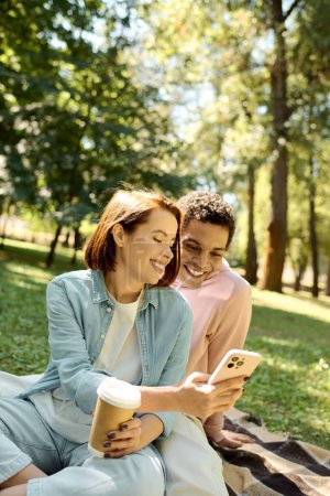 Téléchargez les photos : Couple diversifié en tenue vibrante assis sur une couverture, absorbé dans leur écran de téléphone portable tout en profitant du parc extérieur. - en image libre de droit