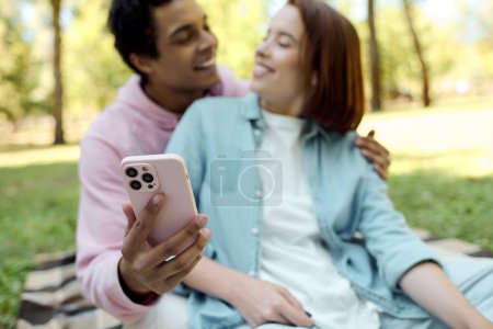 A man and woman in vibrant attire sit on a blanket, holding a cell phone, bonding and sharing a moment in the park.