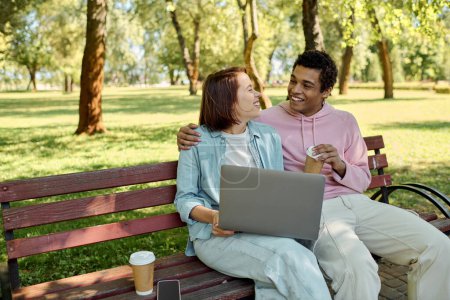 A diverse couple in vibrant attire sits on a park bench, using a laptop together, immersed in their digital world while surrounded by nature.