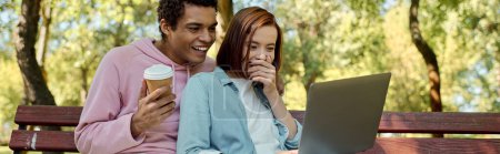Téléchargez les photos : Couple diversifié en tenue vibrante assis sur le banc du parc, absorbé dans l'écran d'ordinateur portable. - en image libre de droit