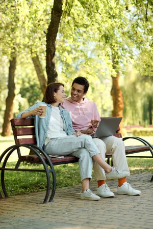 Téléchargez les photos : Un couple élégamment habillé, immergé dans le monde numérique, assis sur un banc de parc, absorbé par l'écran d'un ordinateur portable. - en image libre de droit