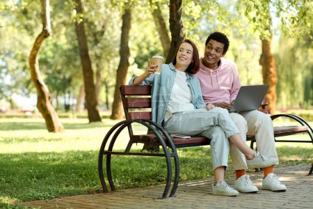 Téléchargez les photos : Un couple diversifié assis sur un banc de parc, travaillant ensemble sur un ordinateur portable, entouré par la nature. - en image libre de droit
