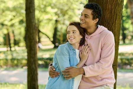 Téléchargez les photos : Un couple aimant et diversifié en tenue vibrante se tient ensemble à côté d'un arbre dans un parc. - en image libre de droit