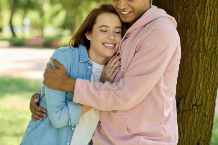 A man and a woman in vibrant attire hug tenderly in front of a majestic tree in a park, showcasing their deep connection. Poster 703329056
