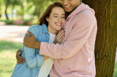 A man and a woman in vibrant attire hug tenderly in front of a majestic tree in a park, showcasing their deep connection. Sweatshirt #703329056