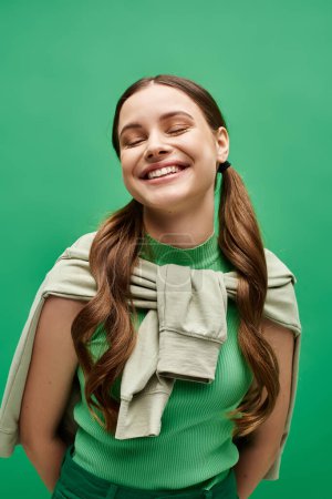 Téléchargez les photos : Une jeune femme heureuse dans la vingtaine aux cheveux longs pose gracieusement dans un pull vert dans un décor de studio. - en image libre de droit