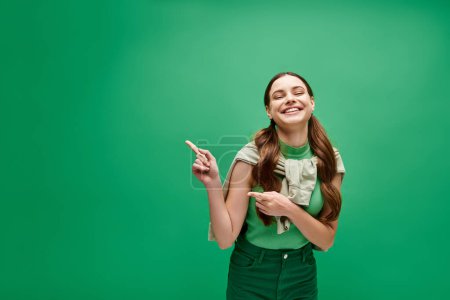 A young woman in her 20s smiles brightly as she points excitedly at something off-camera in a studio setting with a green background.