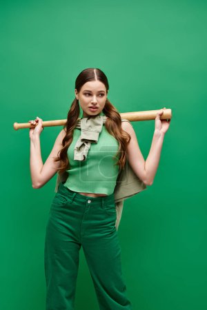 Eine junge Frau in ihren Zwanzigern hält sich einen Baseballschläger über die Schulter und strahlt in einem Studio mit grünem Hintergrund Zuversicht und Gelassenheit aus..
