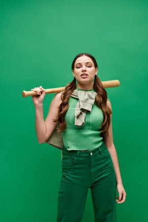 Eine junge Frau in ihren Zwanzigern hält einen Baseballschläger über ihre Schulter in einer selbstbewussten Pose in einem Studio-Setting auf Grün.