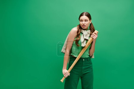 Una joven y hermosa mujer de unos 20 años sostiene con confianza un bate de béisbol frente a un vibrante fondo verde.