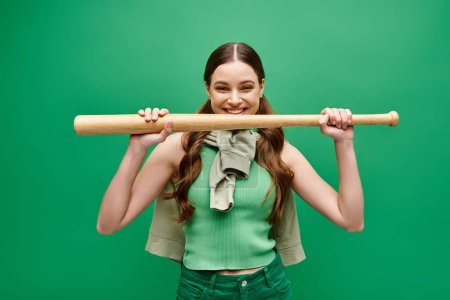 Foto de Una joven y hermosa mujer de unos 20 años sostiene un bate de béisbol sobre su cabeza en una pose dinámica contra un fondo de estudio verde. - Imagen libre de derechos