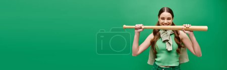 Foto de Una joven y hermosa mujer de unos 20 años sostiene un bate de béisbol frente a su cara en un estudio en verde. - Imagen libre de derechos