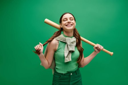 Una joven de unos 20 años sostiene con confianza un bate de béisbol en un estudio sobre un vibrante fondo verde.
