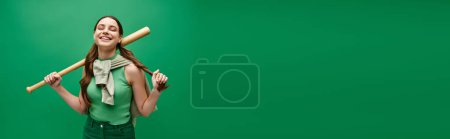 Foto de A young beautiful woman in her 20s confidently holding a baseball bat against a vibrant green background. - Imagen libre de derechos