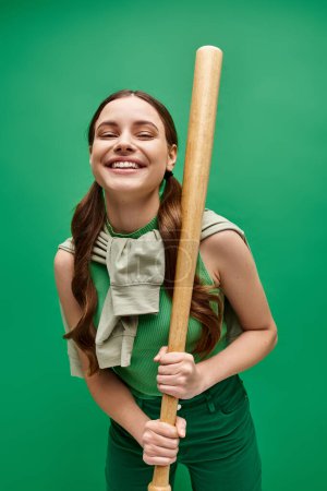 A young woman in her 20s smiles while holding a baseball bat in a studio setting with a green background.