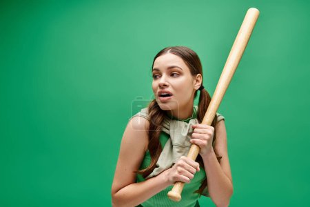 Una joven de unos 20 años sostiene un bate de béisbol con confianza frente a un vibrante fondo verde.
