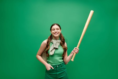 Eine junge Frau in ihren Zwanzigern hält selbstbewusst einen Baseballschläger vor einem auffallend grünen Hintergrund.