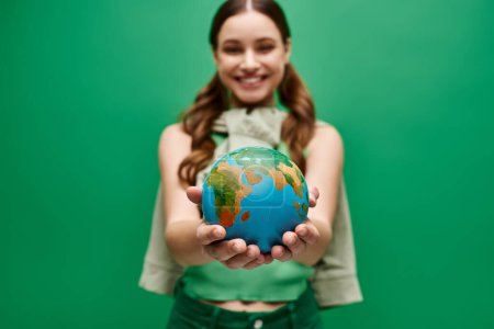 A young woman in her 20s gently holding a small globe in her hands in a studio setting on green background.