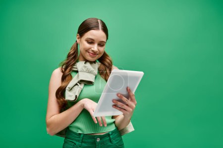 Téléchargez les photos : A stylish woman in her 20s, wearing a green shirt, confidently holds a tablet in a studio setting. - en image libre de droit