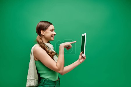Una joven de unos 20 años sostiene una tableta y la señala en un estudio con un fondo verde.