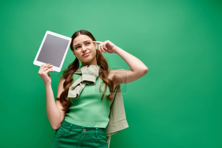 Photo for A young woman in her 20s is holding a tablet and posing confidently in a studio setting with a green background. - Royalty Free Image
