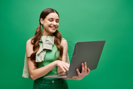 A stylish young woman in a green dress is focused on using a laptop in a modern studio setting.