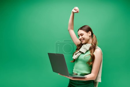 Foto de A young woman in her 20s, wearing a green shirt, confidently holds a laptop in a studio setting. - Imagen libre de derechos