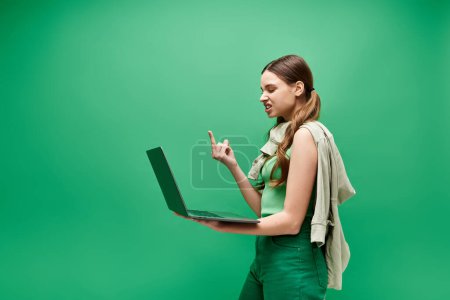 Eine junge Frau in ihren Zwanzigern, in grüner Kleidung, hält einen Laptop in der Hand und zeigt den Mittelfinger in einem Studio-Setting.
