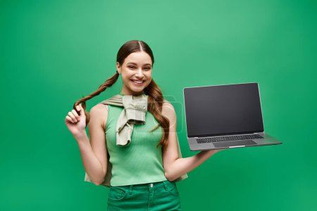A young woman in her 20s confidently holds a laptop in a studio setting with a vibrant green screen background.