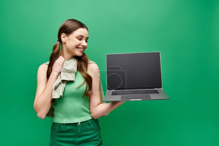 Foto de A young woman in her 20s holding a laptop in front of a vibrant green background. - Imagen libre de derechos