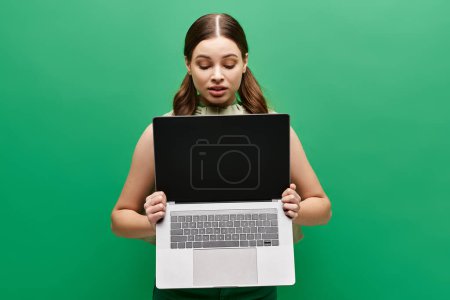 A young woman in her 20s holds a laptop in front of her face, concealing her identity in a studio setting.