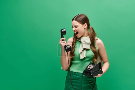 Foto de Young woman in her 20s holding a retro phone, screaming in a studio setting with a green backdrop. - Imagen libre de derechos