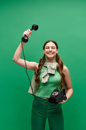 Photo for A young girl in her 20s holding a retro phone in a studio setting on green backdrop. - Royalty Free Image