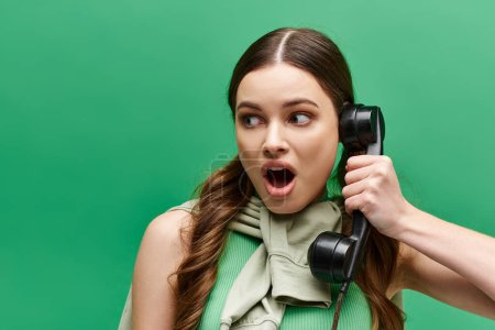 Foto de Una joven sorprendida de unos 20 años sostiene un teléfono retro en su oído, escuchando atentamente. - Imagen libre de derechos