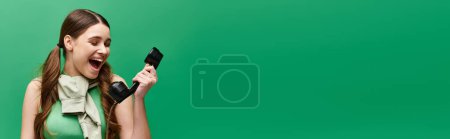 Téléchargez les photos : A young woman in her 20s holds a retro phone, laughing heartily in a studio setting against a green backdrop. - en image libre de droit