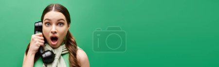 Photo for Shocked woman in her 20s holding a phone to her face, engaged in a phone call, in a studio setting with a green background. - Royalty Free Image