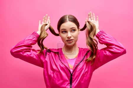 A stylish young woman in her 20s wearing a pink jacket holds her hair in the air against a vibrant pink background.