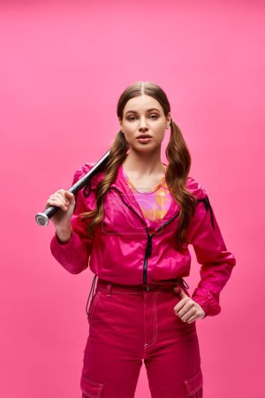 Una chica con estilo de unos 20 años, vestida con un atuendo rosa, sostiene con confianza un bate de béisbol en un estudio con un fondo rosa.