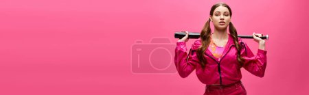 A stylish young woman in her 20s holding baseball bat in front of her face in a studio with a pink background.