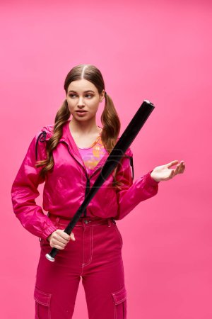 A young stylish woman in her 20s wearing a pink outfit gracefully holding a black baseball bat in a studio with a pink background.