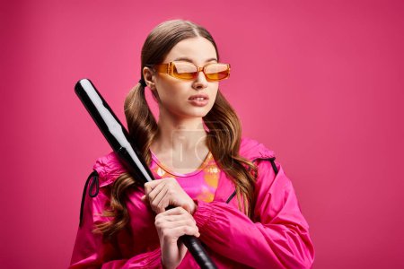 Une femme élégante dans la vingtaine, vêtue d'une veste rose, tient avec confiance une batte de baseball sur un fond rose vif.