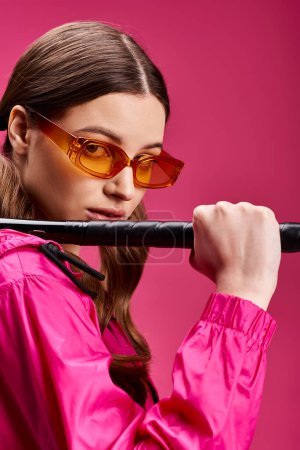 A stylish young woman in her 20s wearing a pink jacket confidently holds a baseball bat against a vibrant pink background.